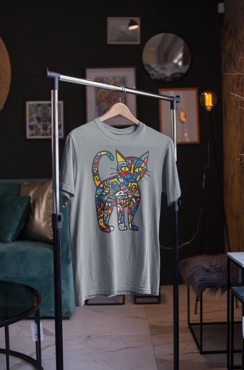 May include: A gray t-shirt featuring a colorful cat design. The cat illustration is filled with abstract shapes in yellow, blue, red, and green. The shirt is hanging on a clothing rack in a room with dark green walls.