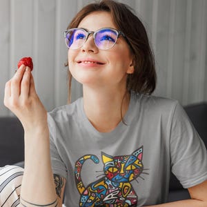May include: A light gray t-shirt featuring a colorful cat design. The cat illustration is filled with various shapes and colors. The person in the image is wearing glasses and holding a strawberry.