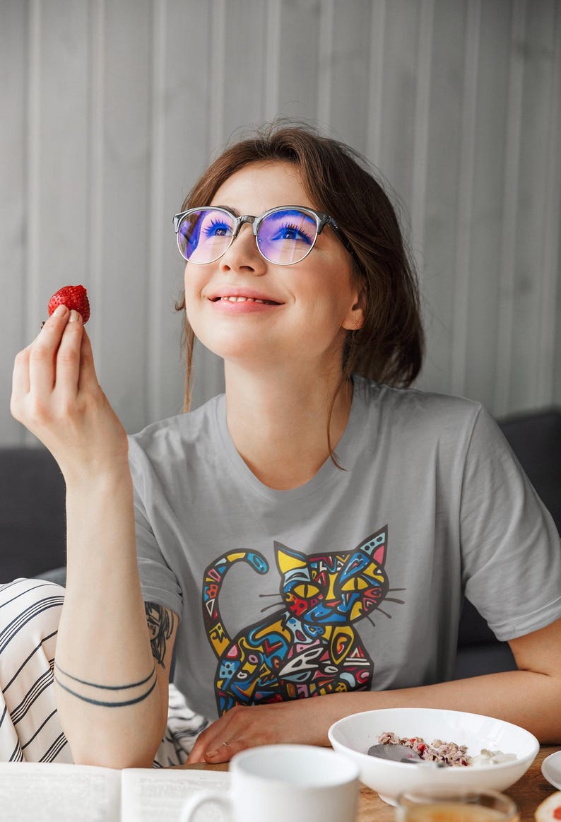 May include: A light gray t-shirt featuring a colorful cat design. The cat illustration is filled with various shapes and colors. The person in the image is wearing glasses and holding a strawberry.