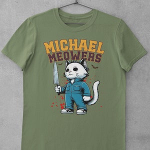 May include: A green t-shirt with a white cat holding a knife. The cat is wearing blue overalls and has a white mask. The text "MICHAEL MEOWERS" is printed in yellow above the cat.