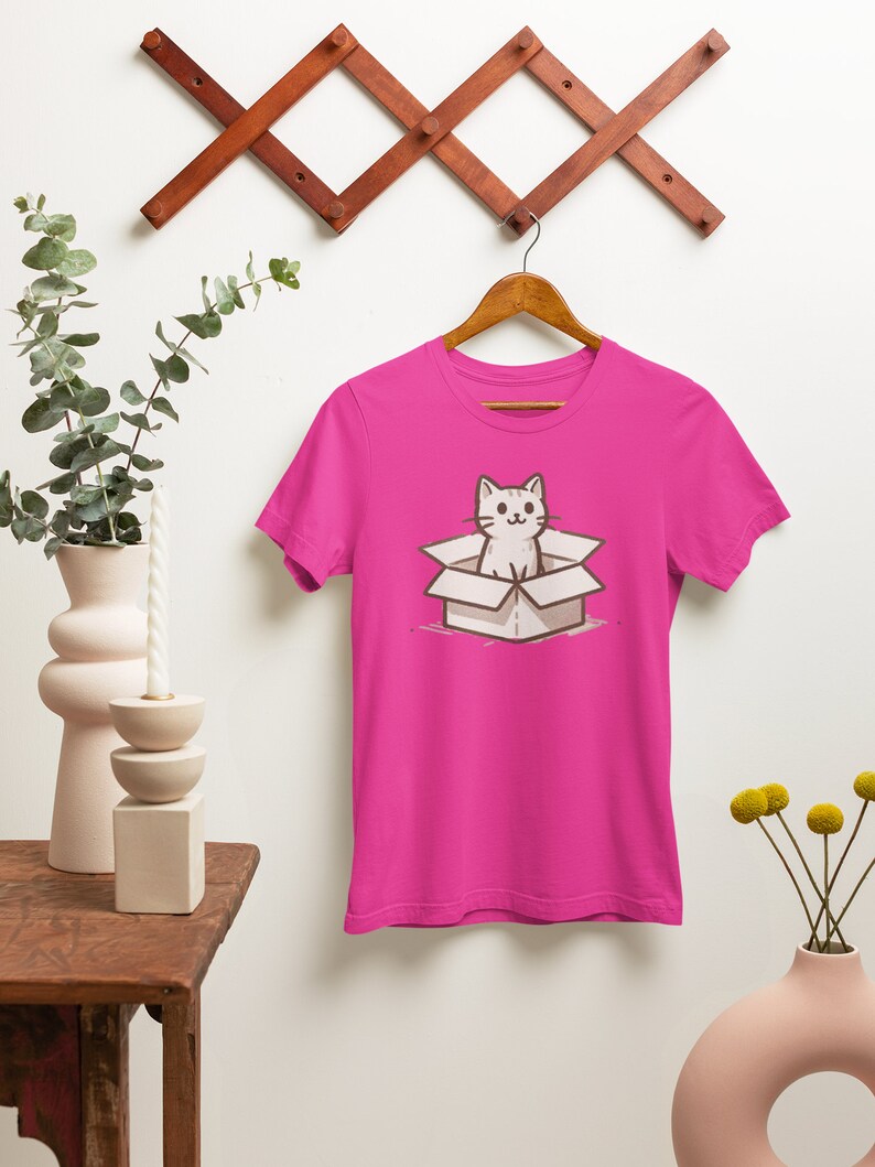 May include: A pink t-shirt with a cartoon illustration of a white cat sitting in a cardboard box.