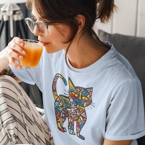 May include: Light blue t-shirt featuring a colorful cat design. The cat illustration is filled with various shapes and patterns in yellow, red, blue, and green. The person is wearing glasses and drinking from a glass.