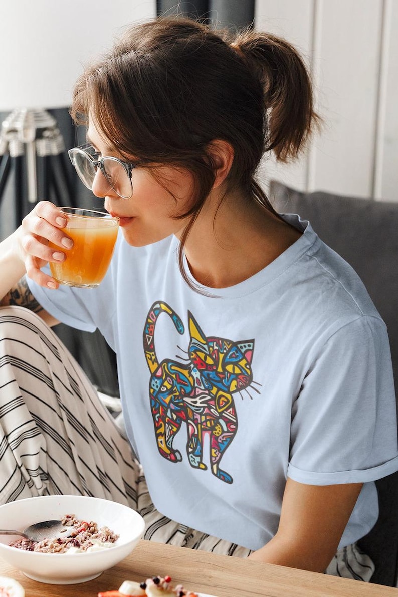 May include: Light blue t-shirt featuring a colorful cat design. The cat illustration is filled with various shapes and patterns in yellow, red, blue, and green. The person is wearing glasses and drinking from a glass.