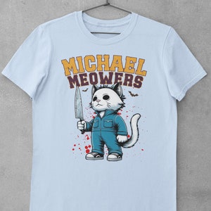 May include: Light blue t-shirt with a cartoon illustration of a white cat holding a knife. The cat is wearing blue overalls and has a white face with black eyes. The text "MICHAEL MEOWERS" is printed in yellow and purple letters above the cat.