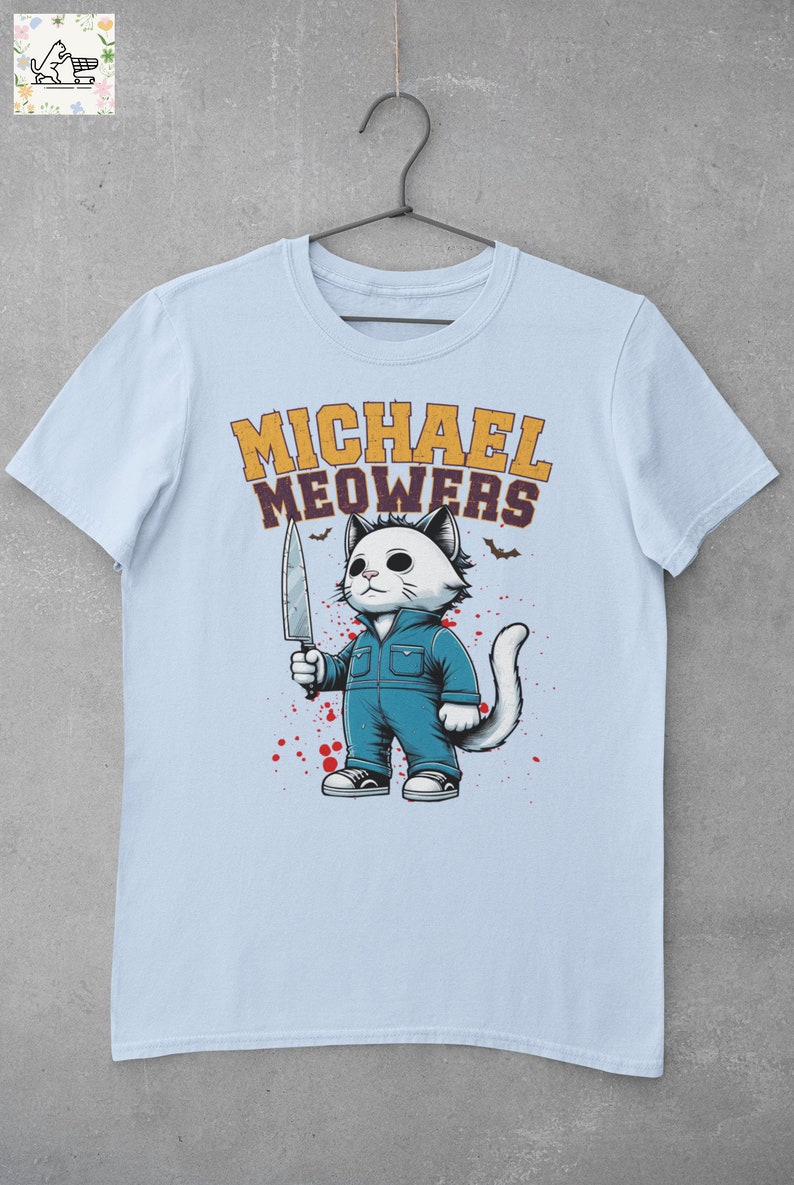 May include: Light blue t-shirt with a cartoon illustration of a white cat holding a knife. The cat is wearing blue overalls and has a white face with black eyes. The text "MICHAEL MEOWERS" is printed in yellow and purple letters above the cat.