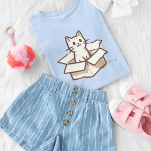 May include: A light blue t-shirt with a cartoon cat in a box graphic. The shirt is paired with blue and white striped denim shorts and pink polka dot canvas shoes.