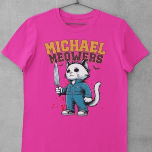 May include: A pink t-shirt with a graphic of a white cat holding a knife. The cat is wearing blue overalls and has a white tail. The text "MICHAEL MEOWERS" is printed in yellow above the cat.