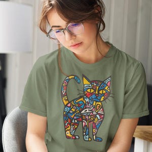 May include: Olive green t-shirt featuring a colorful, abstract cat design. The cat illustration is filled with various shapes and colors, including blue, yellow, red, and orange. The shirt is a short-sleeved crew neck.