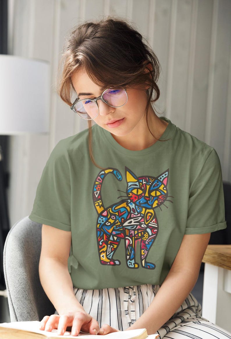 May include: Olive green t-shirt featuring a colorful, abstract cat design. The cat illustration is filled with various shapes and colors, including blue, yellow, red, and orange. The shirt is a short-sleeved crew neck.