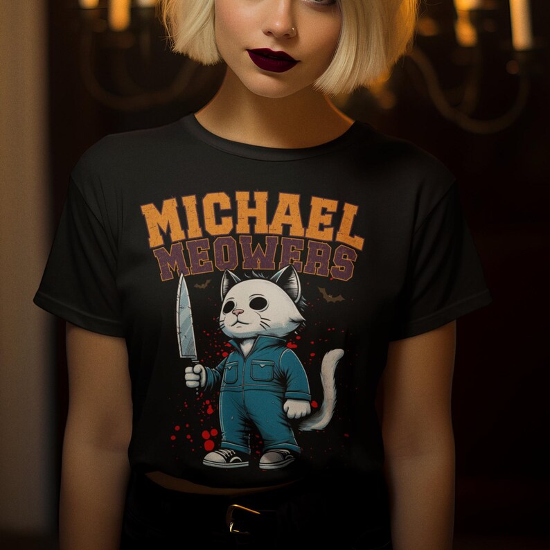 May include: Black t-shirt with a white cat holding a knife. The cat is wearing blue overalls and has a white face. The text "MICHAEL MEOWERS" is printed on the shirt.
