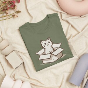 May include: A green t-shirt with a cartoon illustration of a white cat sitting in a brown cardboard box. The cat is looking at the viewer with a happy expression.