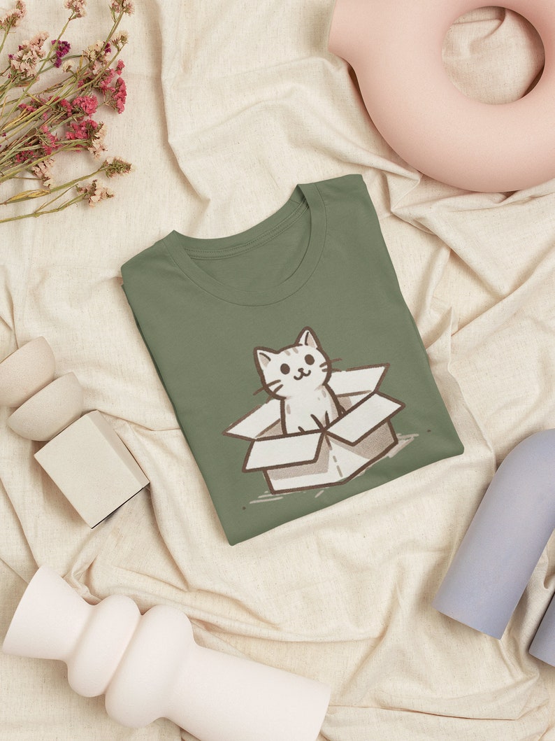 May include: A green t-shirt with a cartoon illustration of a white cat sitting in a brown cardboard box. The cat is looking at the viewer with a happy expression.