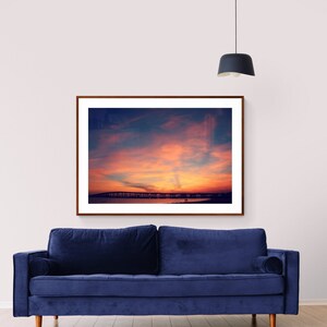 May include: A framed photograph of a sunset over a beach with a bridge in the distance. The sky is a vibrant orange and pink, and the water is a deep blue. The photo is hanging on a white wall above a blue velvet sofa.