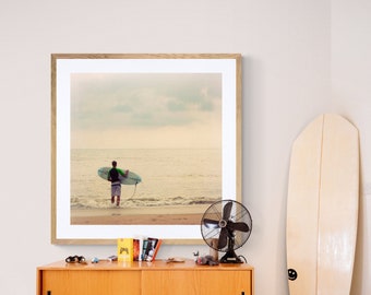 Puede incluir: Una fotografía enmarcada de una persona caminando por una playa con una tabla de surf. La foto está en un marco de madera claro y está colgada en una pared blanca. Hay un armario de madera con dos puertas y un ventilador en primer plano.