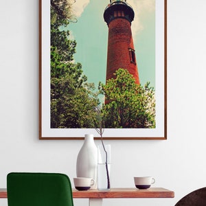 May include: A framed photograph of a red brick lighthouse with a black roof and a white window. The lighthouse is surrounded by green trees and a blue sky with white clouds.