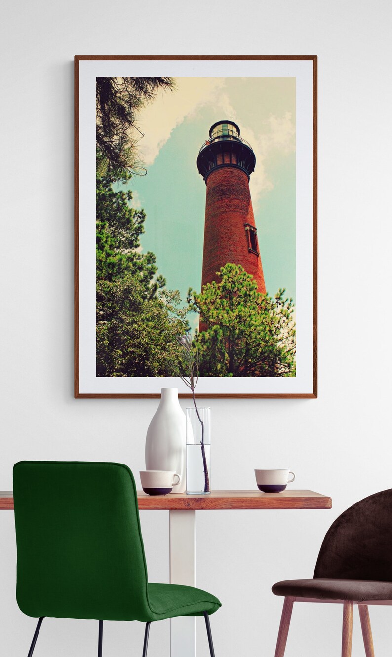 May include: A framed photograph of a red brick lighthouse with a black roof and a white window. The lighthouse is surrounded by green trees and a blue sky with white clouds.