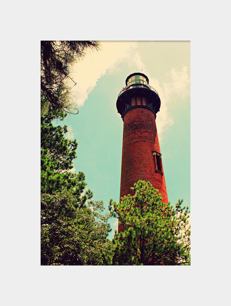 May include: A red brick lighthouse with a black roof and a white lantern room. The lighthouse is surrounded by green trees and a blue sky with white clouds.