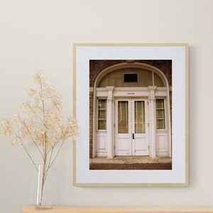 May include: A framed photograph of a white double door with a brick archway above. The door has a transom window and is surrounded by white columns.