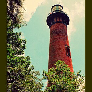 May include: A red brick lighthouse with a black metal roof and a small window. The lighthouse is surrounded by green trees and a blue sky with white clouds.
