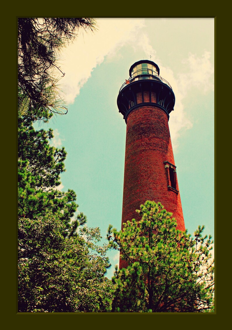 May include: A red brick lighthouse with a black metal roof and a small window. The lighthouse is surrounded by green trees and a blue sky with white clouds.
