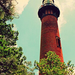 May include: A red brick lighthouse with a black metal roof and a small window. The lighthouse is surrounded by green trees.