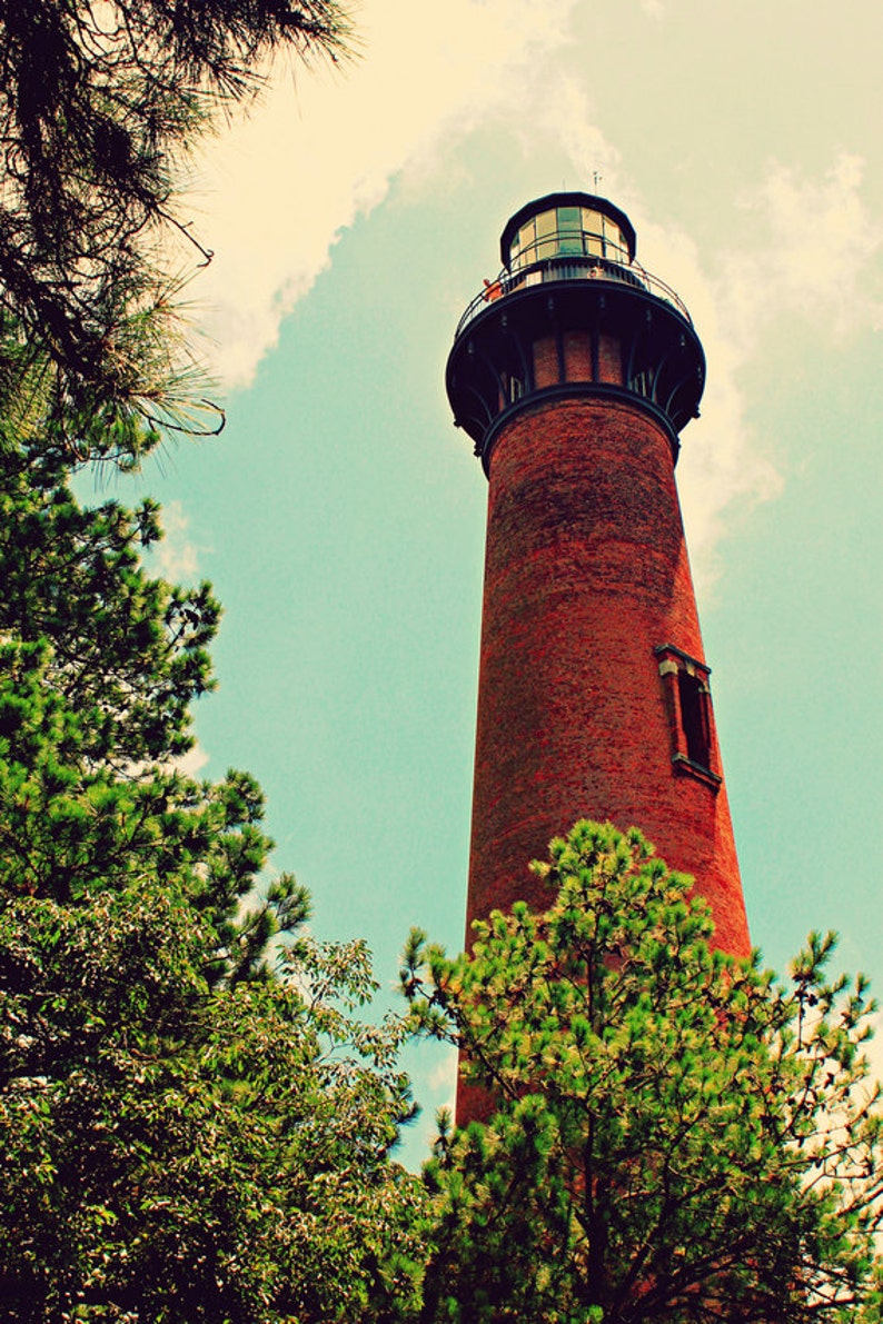 May include: A red brick lighthouse with a black metal roof and a small window. The lighthouse is surrounded by green trees.