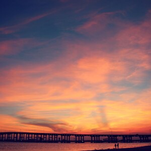 May include: A silhouette of a bridge against a vibrant orange and pink sunset sky. The sky is filled with wispy clouds, creating a dramatic and colorful backdrop.