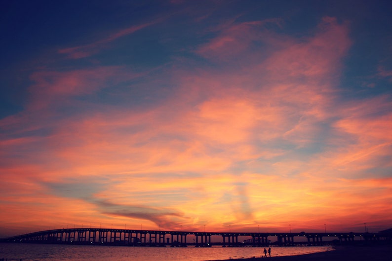 May include: A silhouette of a bridge against a vibrant orange and pink sunset sky. The sky is filled with wispy clouds, creating a dramatic and colorful backdrop.