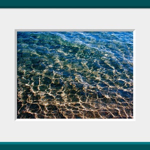 Pu&ograve; includere: Primo piano di acqua blu limpida con la luce del sole che si riflette sulla superficie, creando un effetto scintillante. L'acqua &egrave; bassa, rivelando un fondo sabbioso scuro.