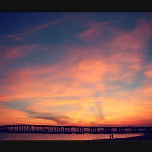 May include: A scenic view of a bridge over a body of water at sunset. The sky is a vibrant orange and pink, with wispy clouds. The bridge is silhouetted against the sky, and there are two people walking on the beach in the foreground.