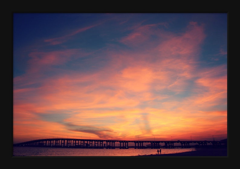 May include: A scenic view of a bridge over a body of water at sunset. The sky is a vibrant orange and pink, with wispy clouds. The bridge is silhouetted against the sky, and there are two people walking on the beach in the foreground.
