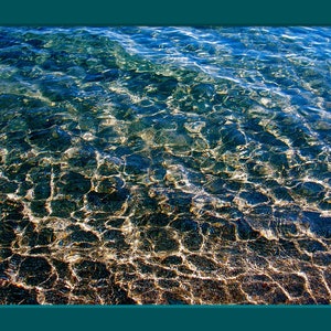 Pu&ograve; includere: Primo piano di acqua blu limpida con un fondo sabbioso. L'acqua &egrave; bassa e la sabbia &egrave; visibile attraverso l'acqua. La luce brilla attraverso l'acqua, creando un effetto scintillante.