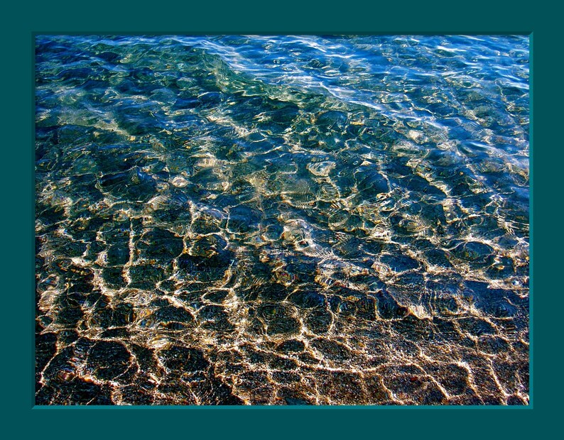 Pu&ograve; includere: Primo piano di acqua blu limpida con un fondo sabbioso. L'acqua &egrave; bassa e la sabbia &egrave; visibile attraverso l'acqua. La luce brilla attraverso l'acqua, creando un effetto scintillante.