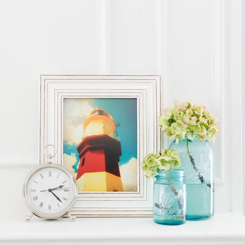 May include: A white picture frame with a lighthouse photograph inside. Two blue glass jars with green flowers are on a white surface.