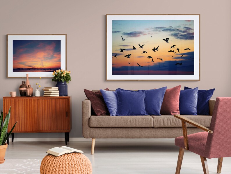 May include: A living room interior with a brown sofa, two blue pillows, a red pillow, and a red armchair. There are two framed pictures on the wall, both depicting a sunset sky with birds flying. A wooden cabinet with a sliding door is in the foreground.
