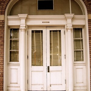 May include: A white double door with a decorative archway above. The door has two panels and a set of blinds on each side. The door is framed by white columns.