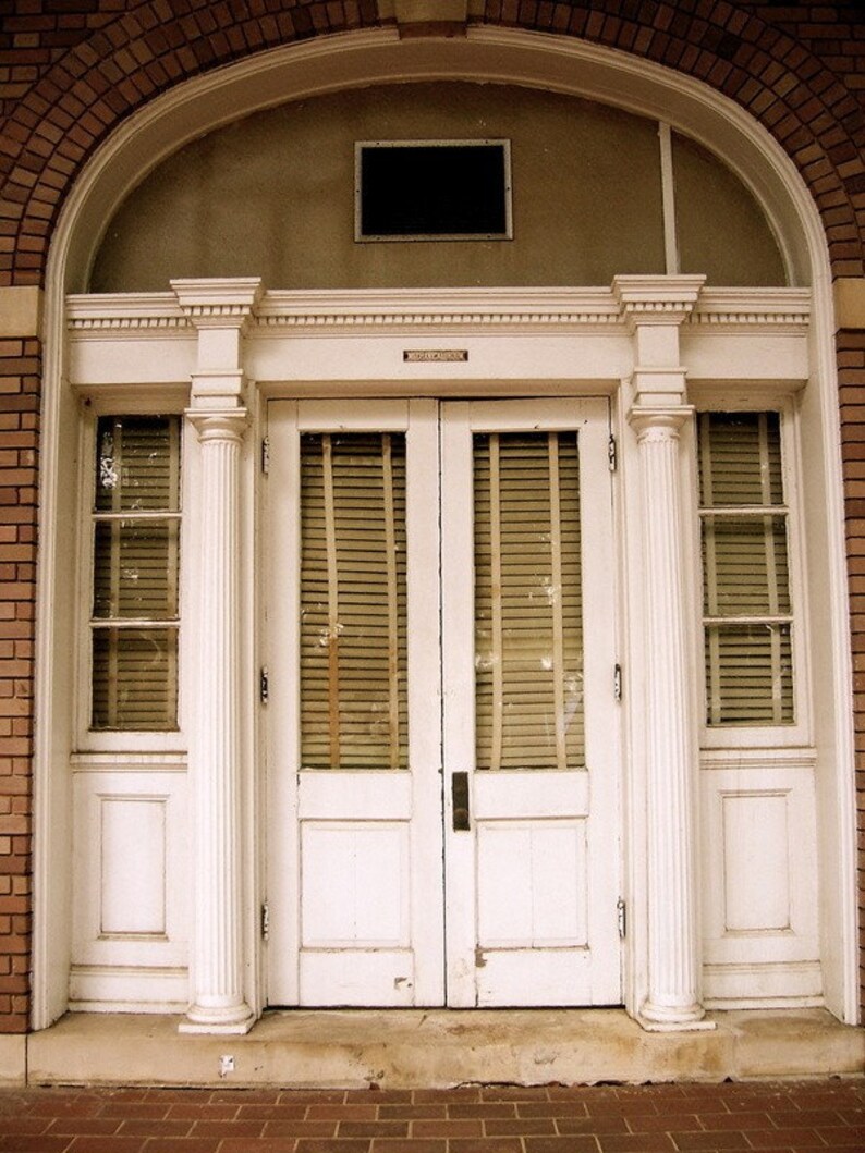 May include: A white double door with a decorative archway above. The door has two panels and a set of blinds on each side. The door is framed by white columns.