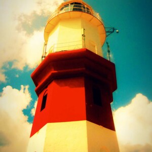 May include: A red and white lighthouse with a white railing and a glass lantern on top. The lighthouse is against a blue sky with white clouds.