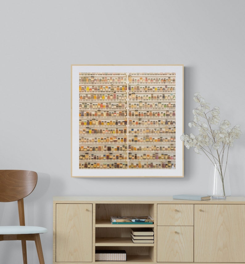May include: A framed photograph of a wall of small glass jars filled with various spices and herbs. The jars are arranged in rows and columns, creating a visually appealing pattern. The photograph is in a light wood frame and is hanging on a white wall.