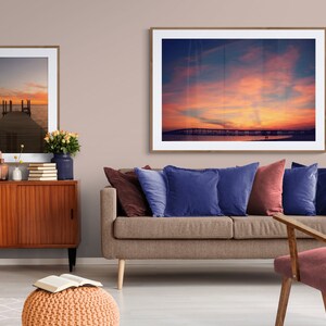 May include: A living room interior with a brown sofa, two blue pillows, a red pillow, a wooden chair, a wooden cabinet, a potted plant, and two framed pictures of sunsets over water.