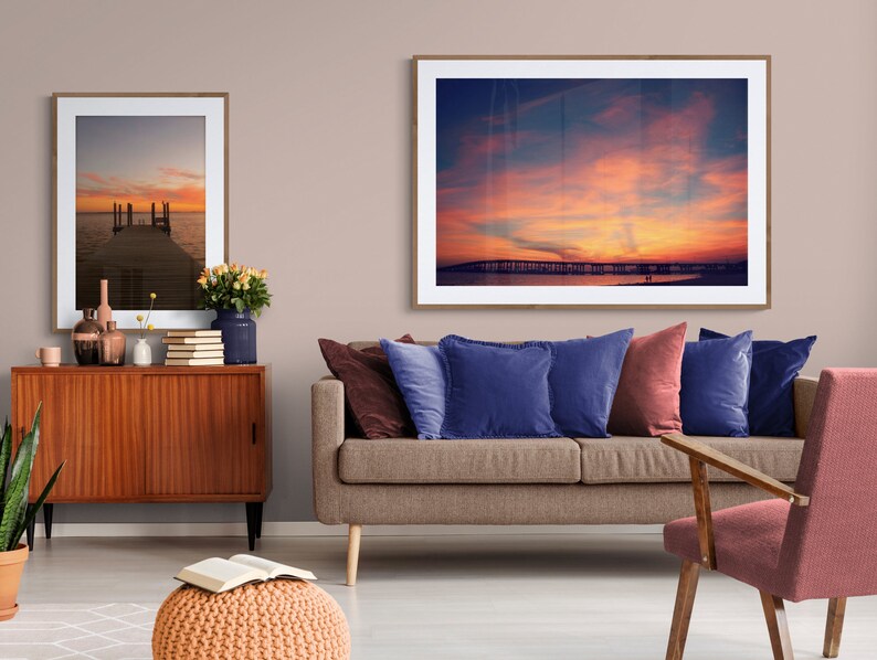 May include: A living room interior with a brown sofa, two blue pillows, a red pillow, a wooden chair, a wooden cabinet, a potted plant, and two framed pictures of sunsets over water.