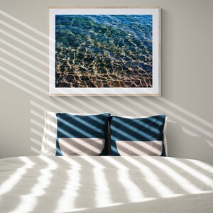 Pu&ograve; includere: Un letto bianco con due cuscini blu e beige. Il letto &egrave; coperto da lenzuola bianche e ha una fotografia incorniciata di acqua blu sopra. La fotografia &egrave; in una cornice di legno.