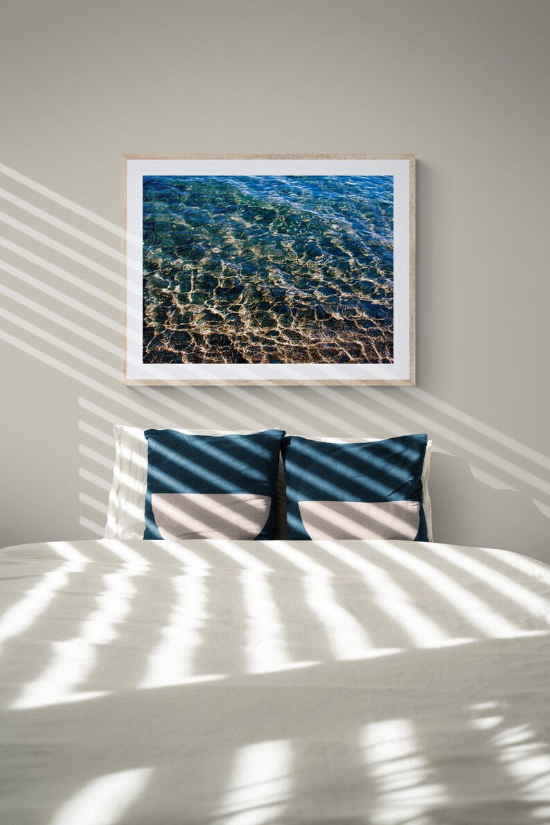 Pu&ograve; includere: Un letto bianco con due cuscini blu e beige. Il letto &egrave; coperto da lenzuola bianche e ha una fotografia incorniciata di acqua blu sopra. La fotografia &egrave; in una cornice di legno.