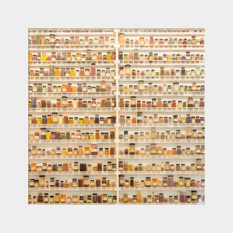 May include: A white shelf unit with rows of small glass jars filled with colorful powders and liquids. The jars are arranged in a grid pattern and are all the same size. The shelf unit is against a white wall.