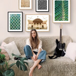 May include: A woman sits on a light brown fuzzy couch with a black electric guitar leaning against the couch. There are five framed pictures on the wall behind the couch. The pictures feature geometric patterns, a building with a large chandelier, and a close-up of a green palm leaf.