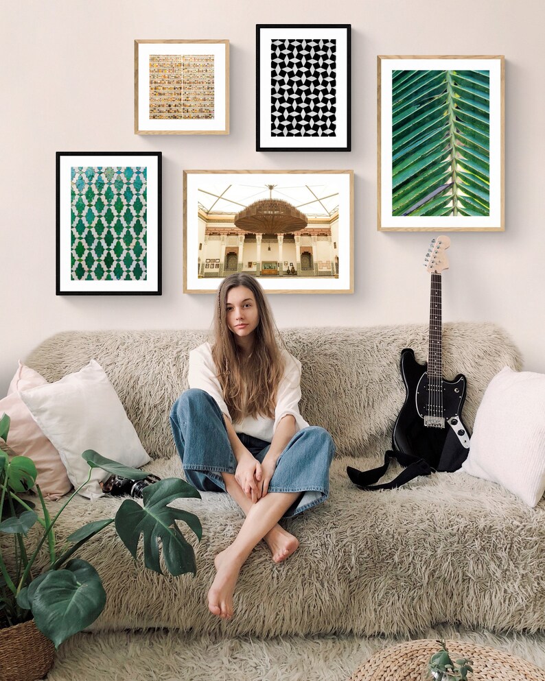 May include: A woman sits on a light brown fuzzy couch with a black electric guitar leaning against the couch. There are five framed pictures on the wall behind the couch. The pictures feature geometric patterns, a building with a large chandelier, and a close-up of a green palm leaf.