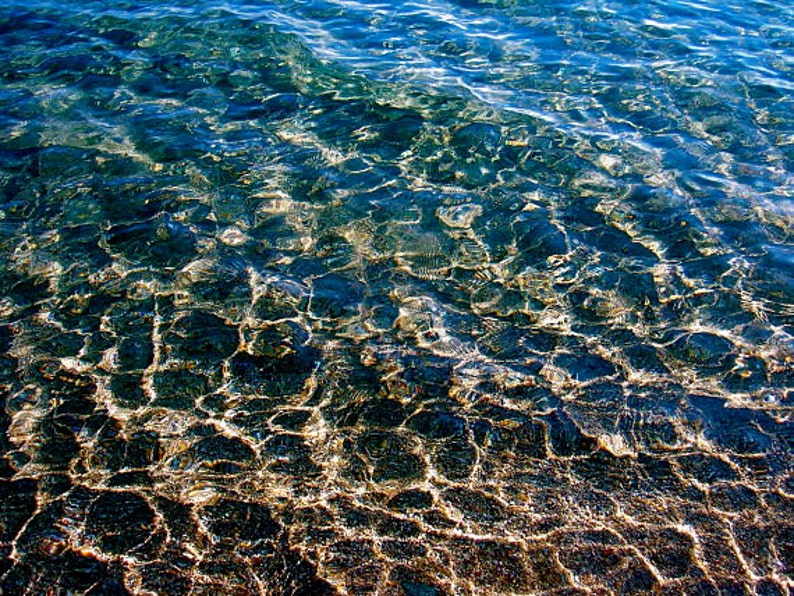 Pu&ograve; includere: Primo piano di acqua blu limpida con la luce del sole che si riflette sulla superficie, creando un effetto scintillante. L'acqua &egrave; bassa, rivelando un fondo sabbioso con un motivo di forme chiare e scure.