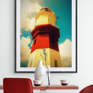 May include: A framed photograph of a red and white lighthouse against a blue sky with white clouds. The lighthouse is tall and has a white roof with a small window. The photograph is in a black frame and is hanging on a white wall.