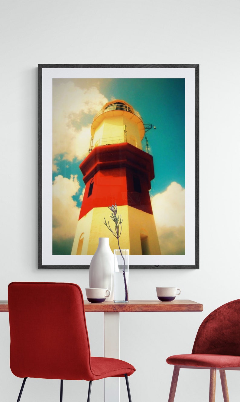 May include: A framed photograph of a red and white lighthouse against a blue sky with white clouds. The lighthouse is tall and has a white roof with a small window. The photograph is in a black frame and is hanging on a white wall.