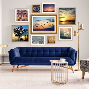 May include: A living room with a blue velvet sofa, a gold metal side table, and a gray patterned armchair. The wall is decorated with a gallery wall of framed photos, including a sunset over the ocean, a cityscape, and a chandelier.